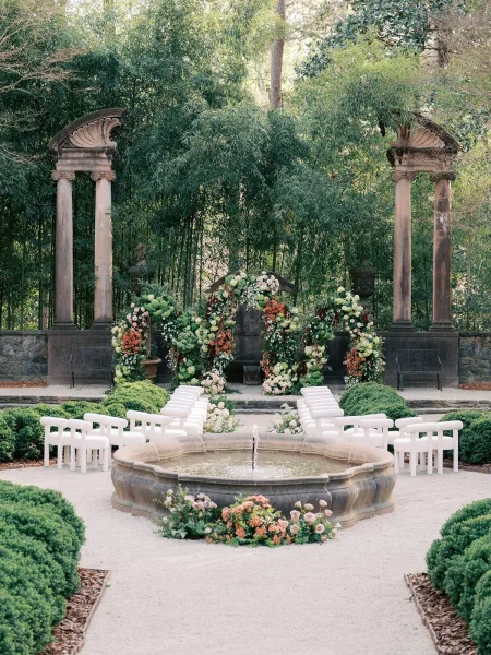 Garden ceremony setup with outdoor ceremony decor centered on a fountain, set in a gravel courtyard beside a stone wall.