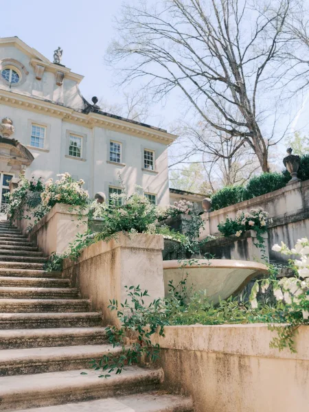 Staircase floral installation draping stone steps, wedding staircase flowers in classic urns, set before a grand mansion facade and hedges.