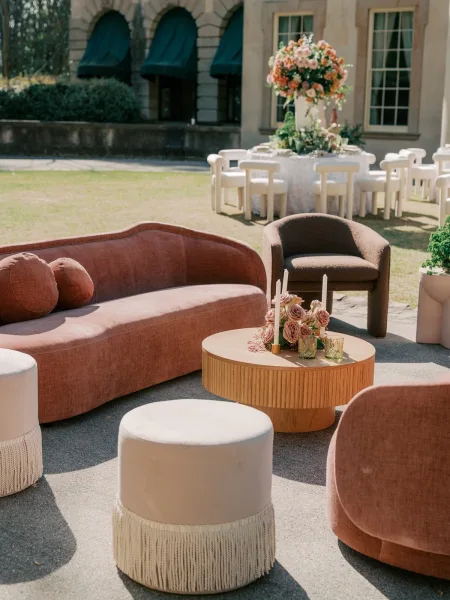 Wedding lounge setup for a cocktail hour lounge featuring fringe ottomans on a lawn beside a stone building facade and garden shrubs.