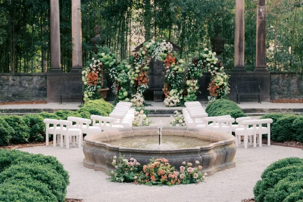 Outdoor ceremony setup for a garden wedding ceremony arranged around a fountain in a gravel courtyard bordered by hedges.