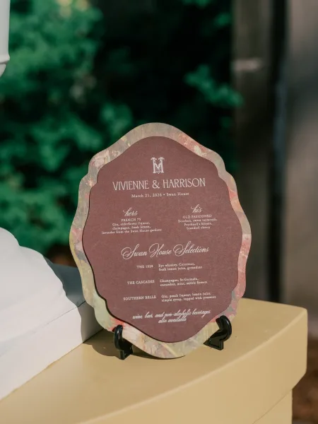 Signature drink sign for the wedding bar menu, featuring a scalloped edge, on a pedestal amid lush garden greenery.
