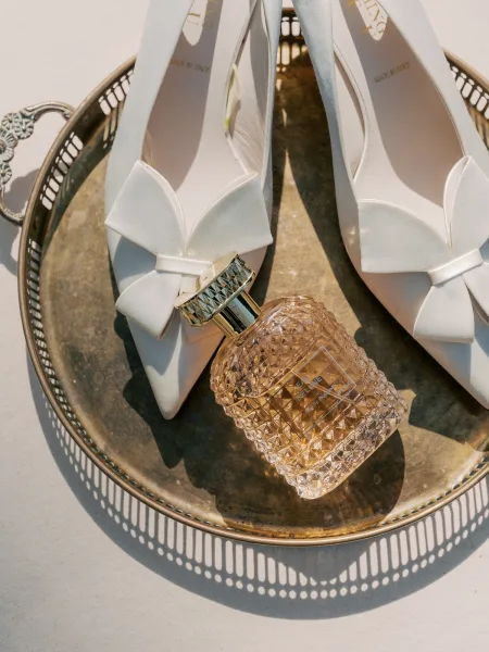 Bridal shoes, delicate bow bridal heels arranged with a perfume bottle on soft linen fabric in a minimalist arrangement.