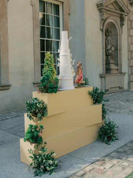 Wedding cake, a tall white cake adorned with sugar leaves, set before a stone wall and window in a cobblestone courtyard.