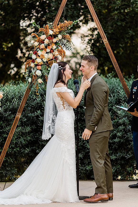 Wedding vows during outdoor wedding ceremony as bride in lace dress and cathedral veil faces groom in olive green suit under floral triangle arch