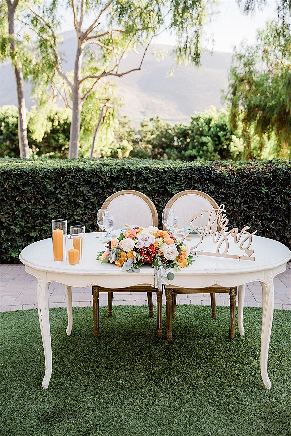 Sweetheart table decor with a white table, round-back chairs, floral centerpiece and wood script sign, set outdoors with mountain views