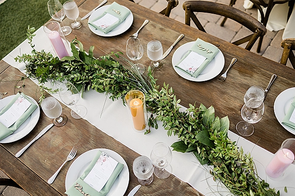 Reception tablescape on a farm table wedding reception setup with white runner, greenery garland, pillar candles, sage napkins on brick patio