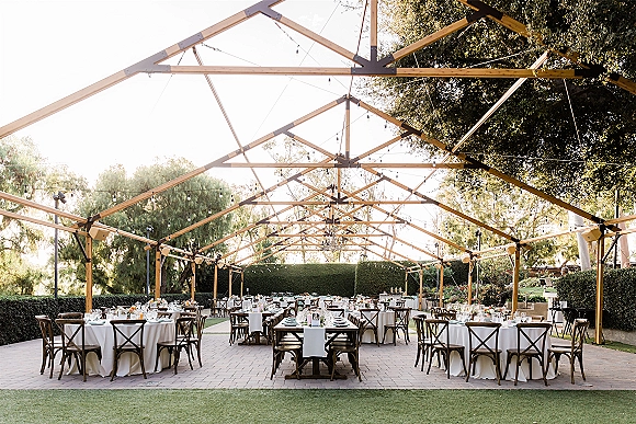 Outdoor reception setup with a clear top tent wedding, string lights, round and banquet tables, white linens, and floral centerpieces on a garden lawn