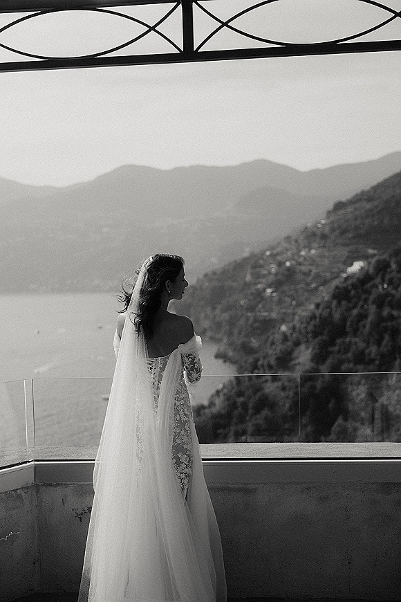 Bridal portrait of bride from behind in lace off-shoulder gown, looking over shoulder with long veil on balcony above lake and mountains