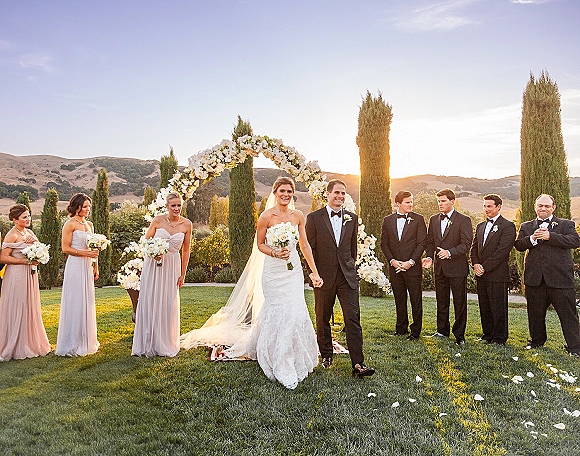 Wedding recessional as bride and groom walk hand in hand under a white floral arch, bouquet and veil glowing in sunset light on a lawn