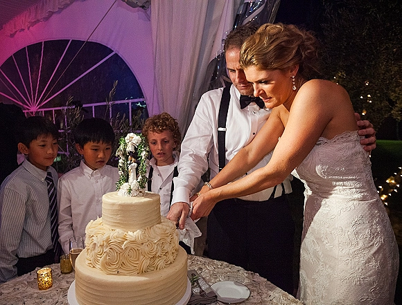 Wedding cake cutting by bride and groom cutting cake beside champagne flutes, under string lights in a draped tent at night with greenery backdrop