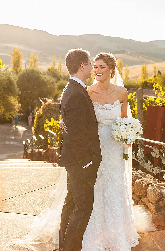 Couple portrait of bride and groom laughing and embracing, her veil and white bouquet glowing at sunset beside a vineyard stone wall