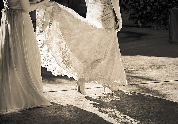 Wedding dress detail with lace wedding dress train lifted to reveal white platform bridal heels on a concrete walkway in sunlight and shadows