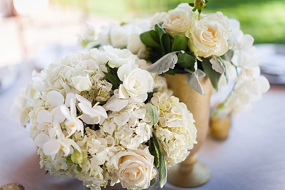 Wedding centerpiece of white floral centerpiece blooms—roses, hydrangeas, and orchids—in a gold vase on linen tablecloth outdoors