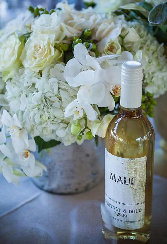 Wedding wine bottle with a custom wine bottle label beside white roses, hydrangeas, and orchids on a reception table with blurred indoor backdrop