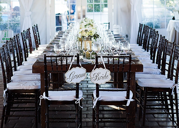Reception tablescape with a long banquet table wedding setup, white florals and greenery garland in a bright, draped tent with windows