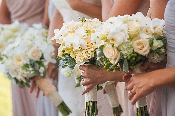 Bridesmaid bouquets with white orchid blooms and cream roses, tied with greenery and berry accents, held in outdoor light