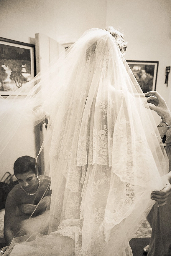 Bride getting ready as a bridesmaid helps with a long tulle wedding veil over a lace dress in a simple indoor room
