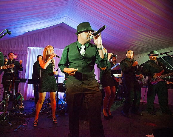 Wedding band performing live wedding music on a stage with microphones, trumpet, drums and bass under a white draped tent with purple uplighting