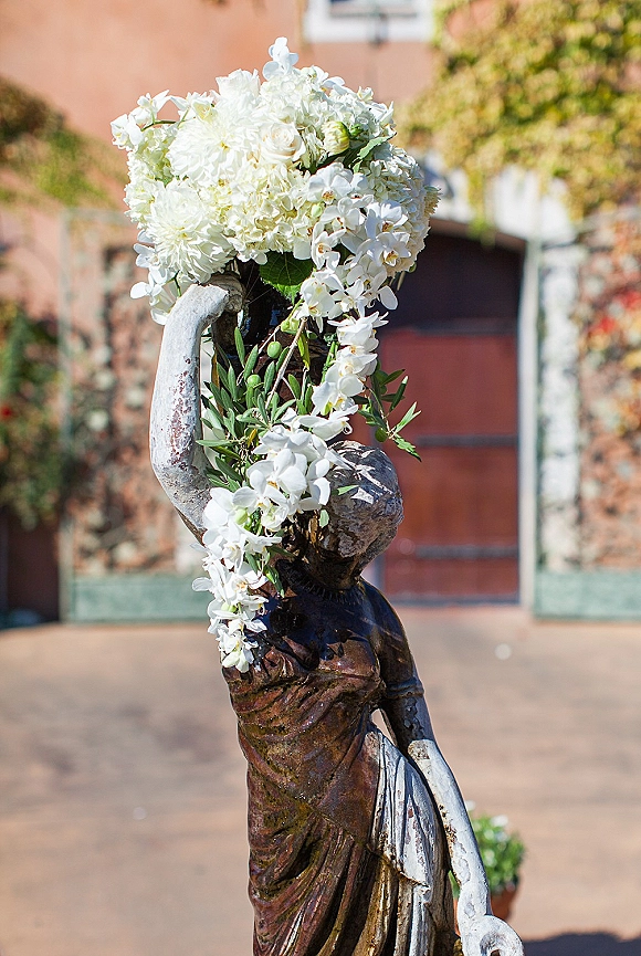 Wedding floral arrangement of white wedding florals with orchids, hydrangea, roses and olive greenery on a statue pedestal in a brick courtyard