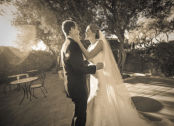 Wedding couple portrait of the bride and groom embrace as she holds his face, her long veil glowing on a sunlit patio with trees behind