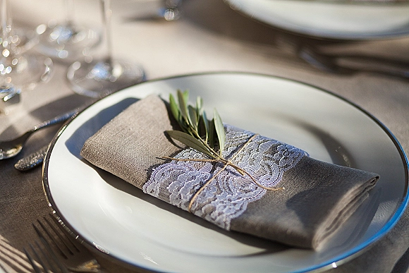 Wedding place setting with a linen napkin fold wrapped in lace and twine, topped with an olive branch beside silverware and wine glass