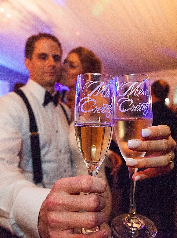 Wedding champagne toast as bride and groom clink custom champagne flutes with etched lettering under tented reception lights, guests blurred behind