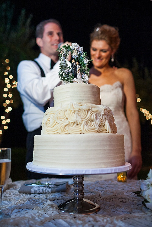 Wedding cake with two-tier rosette frosting and floral wreath topper on a lace table, with bride and groom under string lights at night
