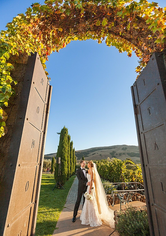 Couple portrait of bride and groom holding hands by open iron gates, her veil and bouquet flowing along a vineyard garden path
