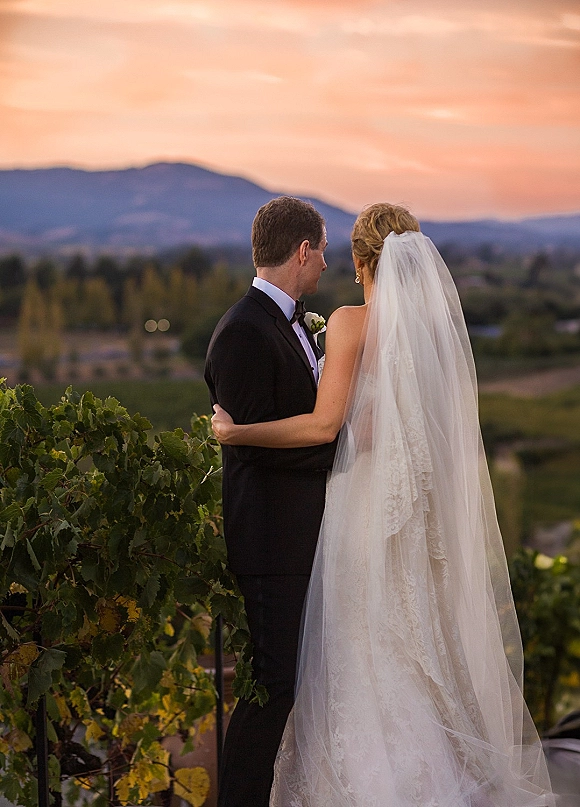 Couple portrait of bride and groom at sunset, seen from behind embracing, her cathedral veil over lace dress above vineyard hills