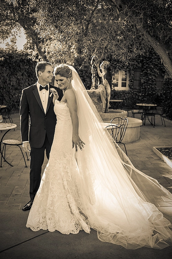 Couple portrait of bride and groom smiling, bride looking down in strapless lace gown and long veil, with ivy courtyard fountain backdrop