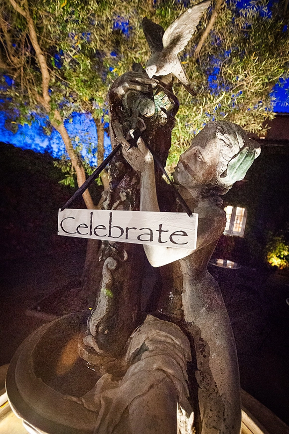 Wedding signage celebrate wedding sign hanging from ribbon on a wooden sign beside a statue, under tree branches in a night courtyard