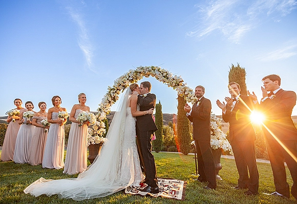 Wedding kiss under a white floral arch as the bride’s veil flows, bridal party lined up on an outdoor lawn at sunset