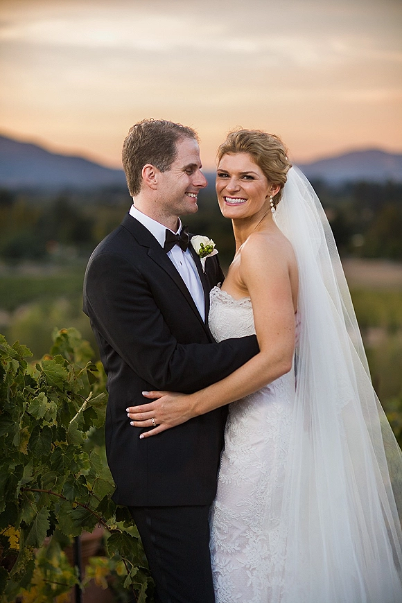 Couple portrait at sunset with bride in strapless lace dress and veil embracing groom in black tuxedo, mountains and vineyard behind