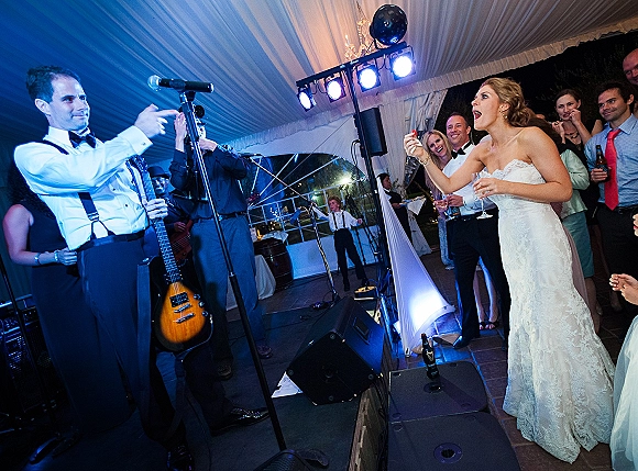 Wedding reception dancing as the bride sings into a microphone beside the groom in suspenders under a draped tent ceiling and stage lights