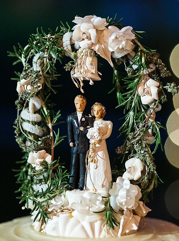 Wedding cake topper with bride and groom cake topper figurines inside a white floral greenery wreath, set against dark bokeh lights