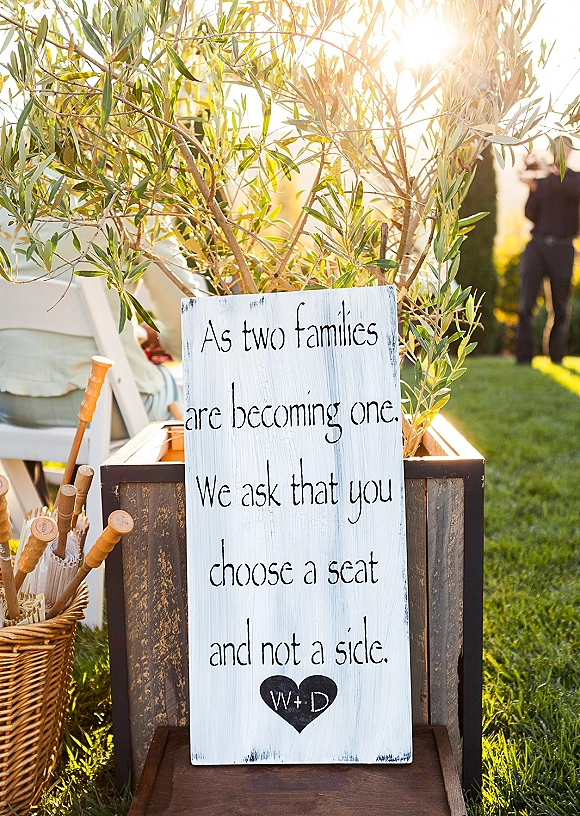 Wedding signage with a choose a seat not a side sign on a wooden crate, olive branches and parasols in a wicker basket on a sunny lawn