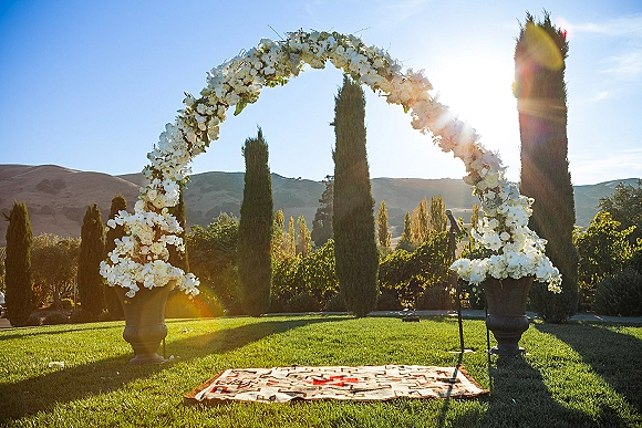 Wedding ceremony arch with white flowers and urn planters on a rugged aisle, set on a vineyard lawn with cypress trees and hills