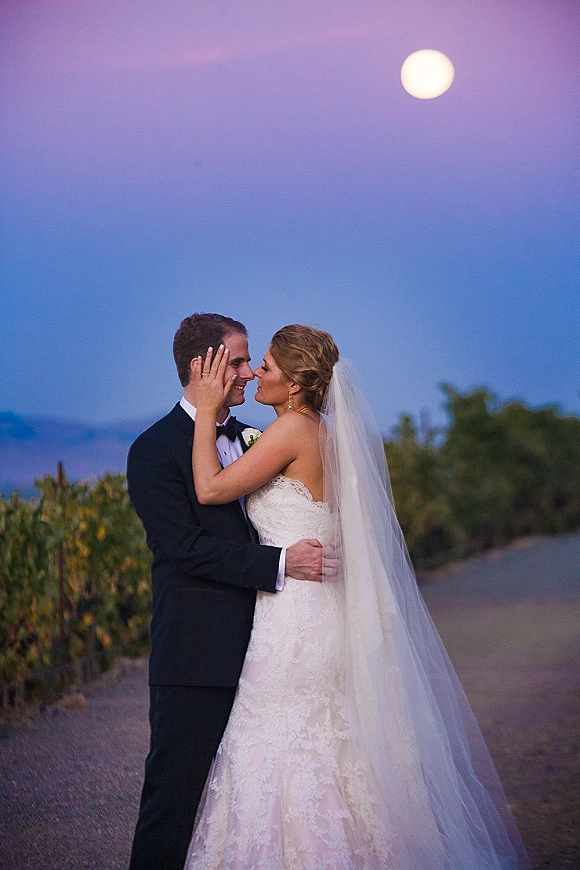 Wedding kiss portrait of bride and groom kissing under a full moon at twilight in a vineyard, her veil flowing as they embrace
