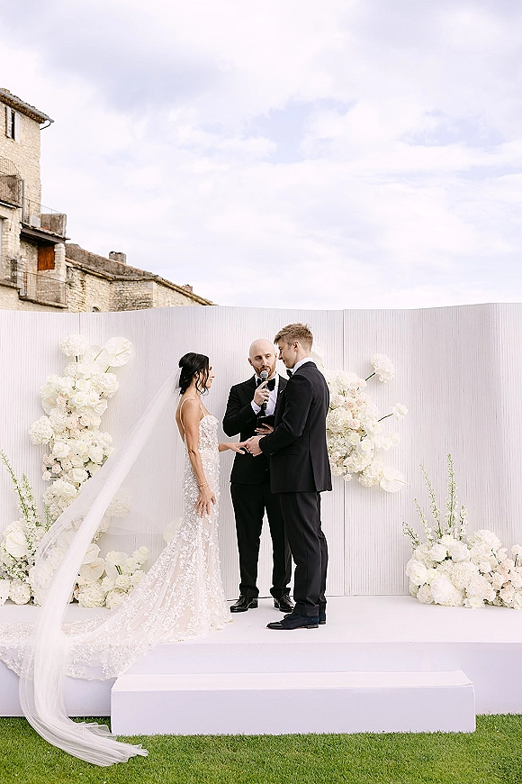 Wedding vows as bride in lace dress and veil holds groom’s hand while officiant speaks on a white floral stage outdoors by stone building