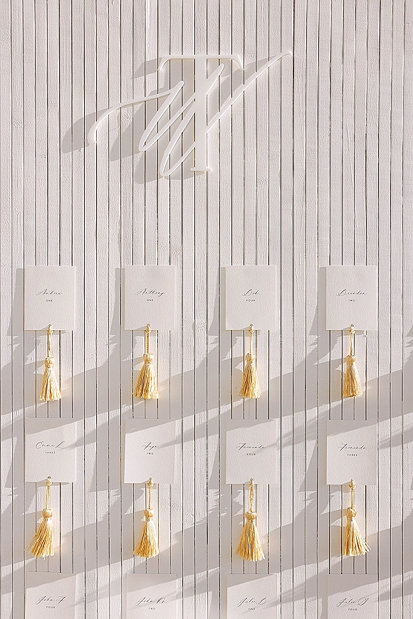 Seating chart display with an escort card wall of calligraphy cards on hooks, gold tassels, and monogram sign against sunlit white panels