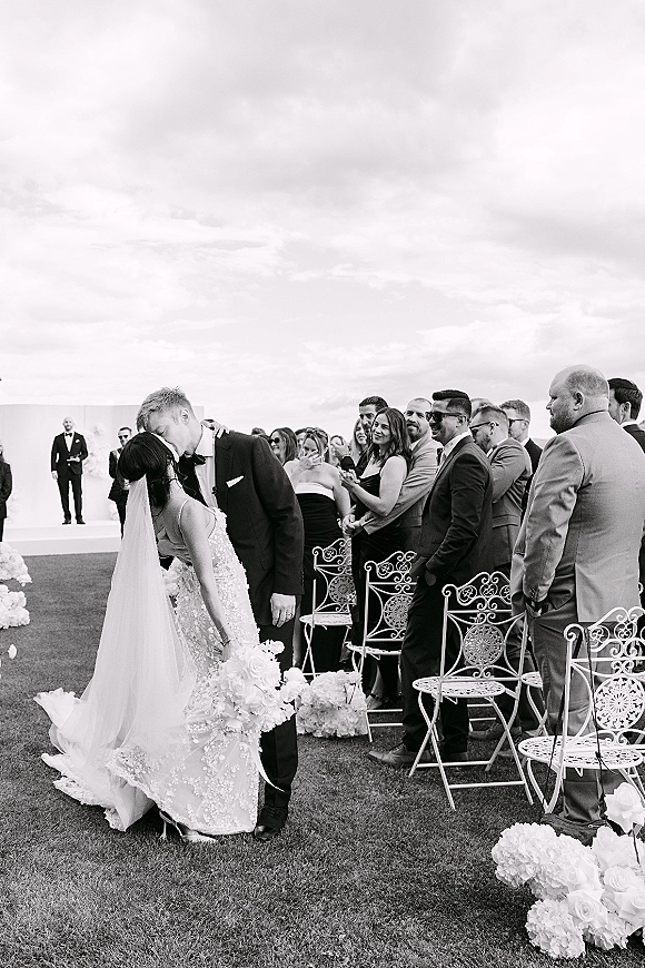 Wedding kiss at an outdoor ceremony as groom dips bride in lace dress and veil, holding bouquet beside floral aisle, guests watching under cloudy sky