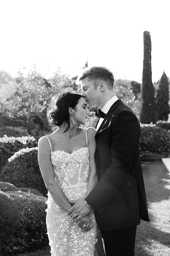 Wedding couple portrait of bride and groom holding hands as he kisses her forehead in a sunlit garden, lace dress and tuxedo visible