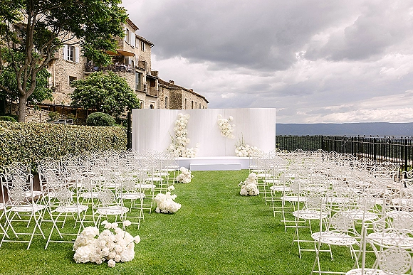Ceremony setup with ornate white metal chairs and floral arrangements, featuring curved white backdrop panels on a lawn by a stone building.