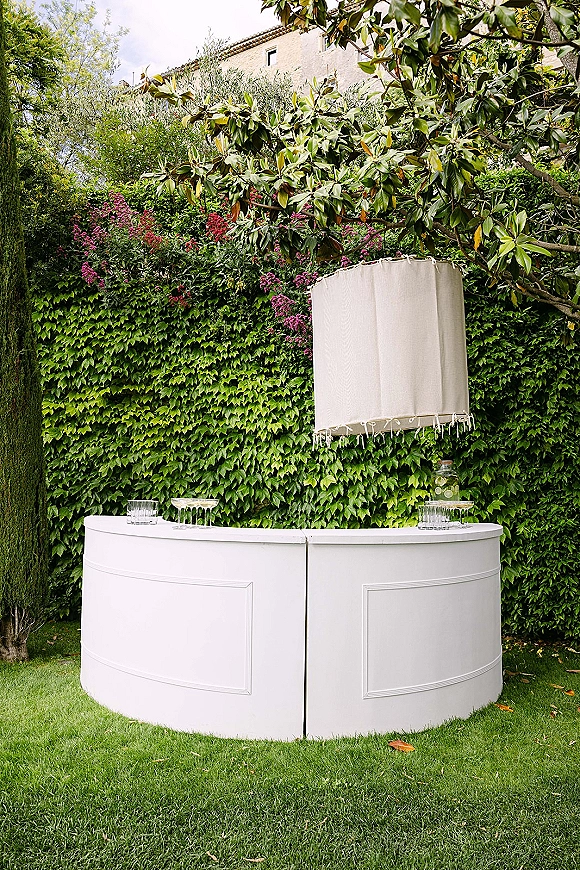 Wedding bar setup with a white curved portable wedding bar, glasses and glass beverage dispenser, set on a garden lawn by an ivy wall