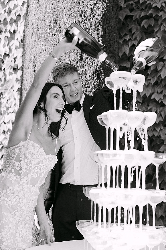 Champagne tower wedding scene as bride in lace dress and groom in tuxedo pour bottles into coupe glasses before an ivy wall backdrop