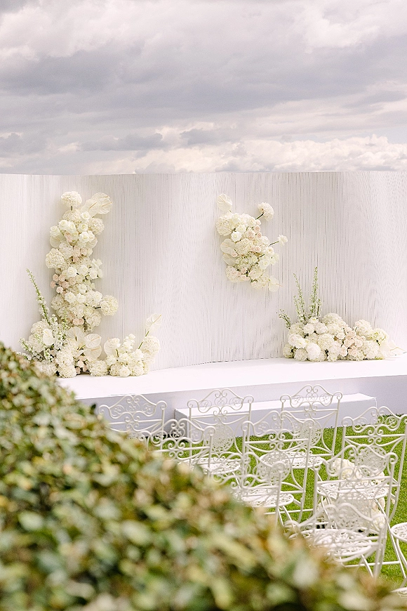 Ceremony backdrop with white floral arrangements framing a white ceremony stage, wrought iron chairs on a lawn under a cloudy sky