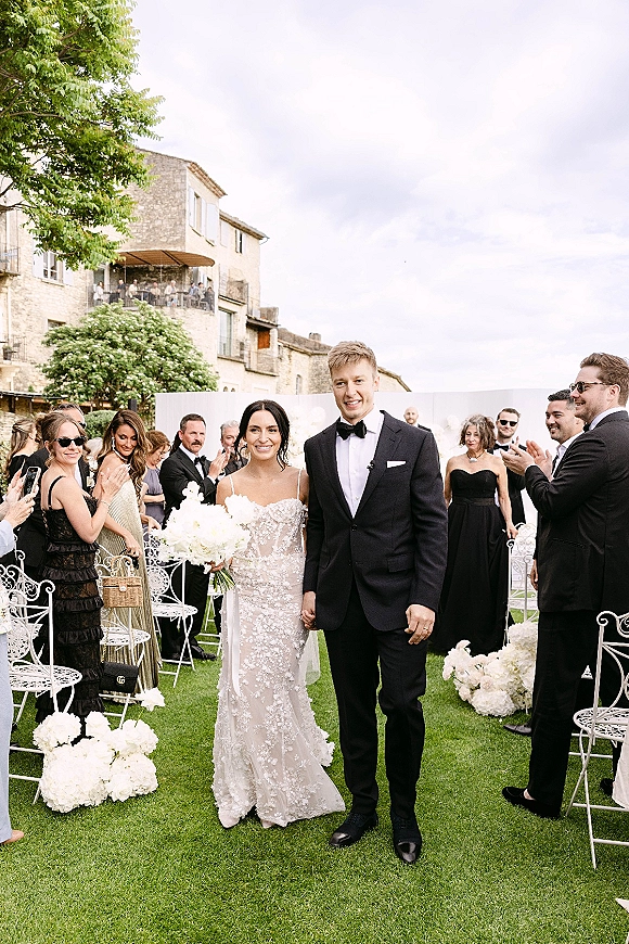 Recessional portrait of bride and groom walking aisle, bouquet raised, in lace gown and black tux as guests clap by stone terrace.