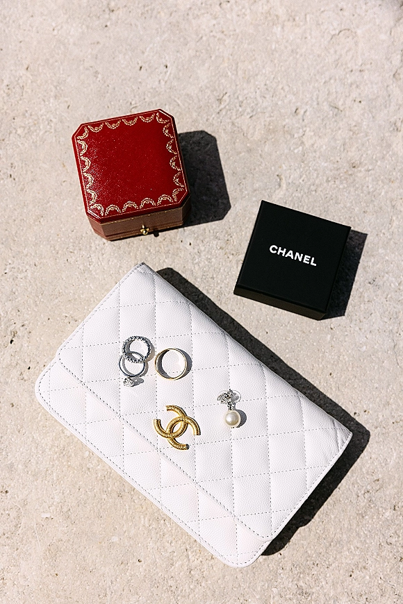 Wedding rings flatlay with engagement ring and band beside pearl drop earrings, quilted white clutch, and ring boxes on stone surface