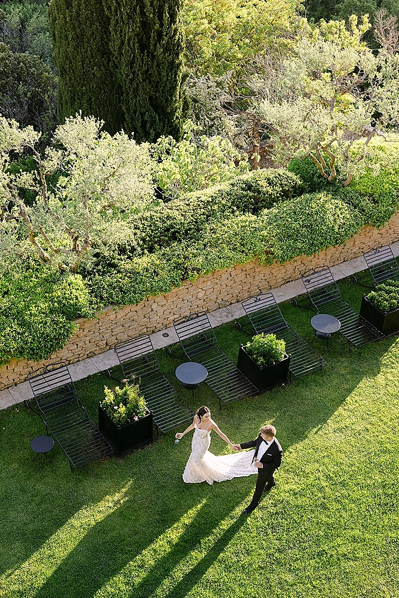Couple portrait of bride and groom holding hands, her veil and dress train flowing as they toast with champagne on a garden lawn