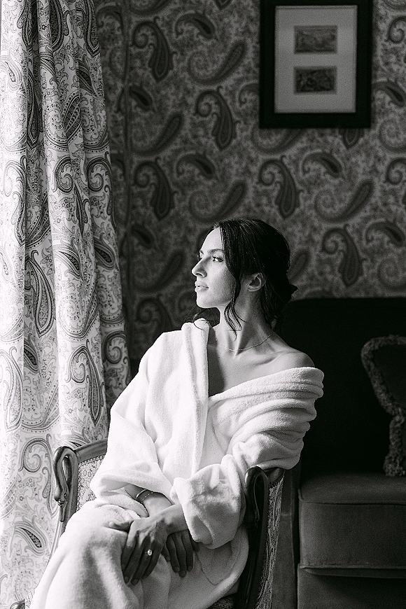 Bridal getting ready portrait of a bride in robe, seated by window light with a necklace, framed artwork, and patterned wallpaper behind her
